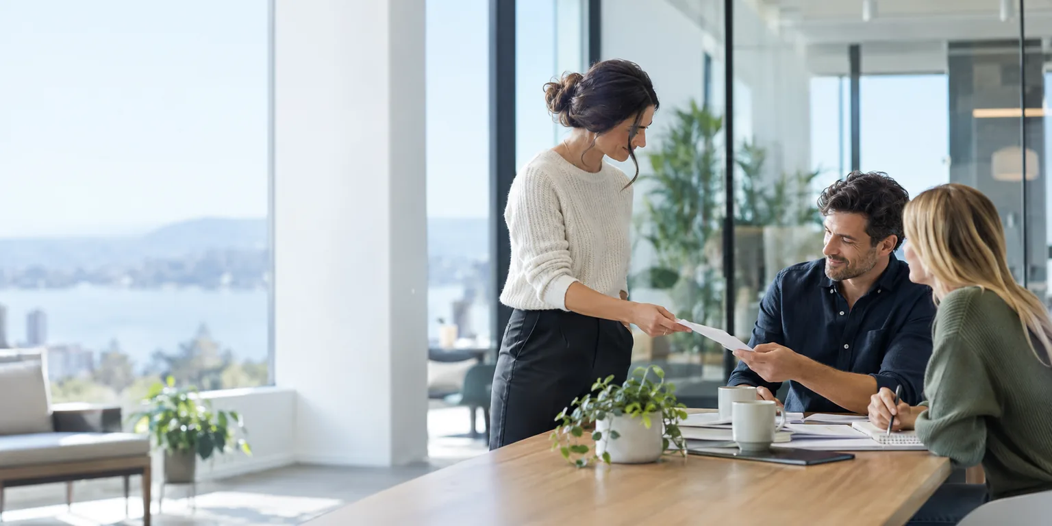 Three colleagues in a bright brokerage workspace, one handing work to another.